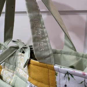 May include: Close-up of a patchwork tote bag with a light green handle embroidered with "06/27/26". The bag features a mix of floral and solid fabric panels in green, yellow, and white, with a quilted design. The bag has a light green interior.