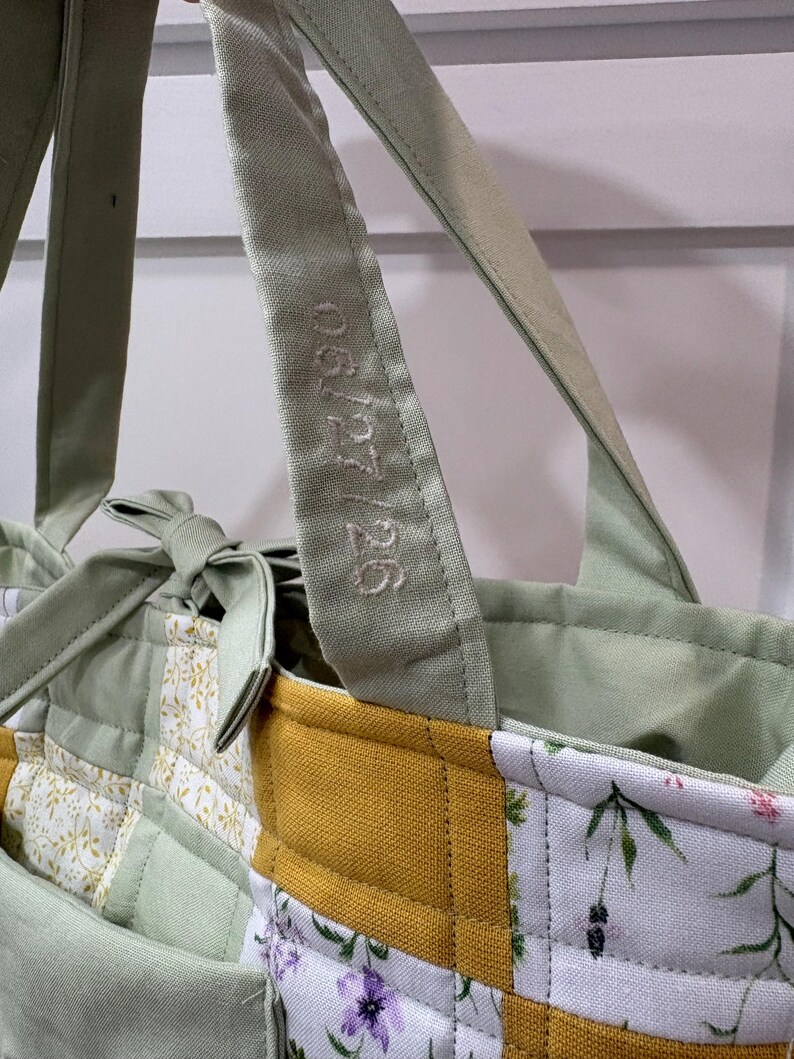 May include: Close-up of a patchwork tote bag with a light green handle embroidered with "06/27/26". The bag features a mix of floral and solid fabric panels in green, yellow, and white, with a quilted design. The bag has a light green interior.