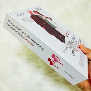May include: A white book titled "Draco Malfoy & the Mortifying Ordeal of Being in Love" with illustrations of characters. The book has red heart-shaped glasses on the side and the text "Isthisselfcare".