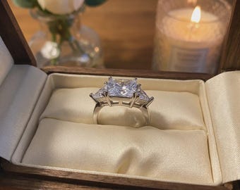 May include: A silver-toned ring featuring a large, square-cut center stone flanked by two smaller triangular-cut stones on each side. The ring is displayed inside a wooden box lined with cream-colored fabric, showcasing a polished metal band.