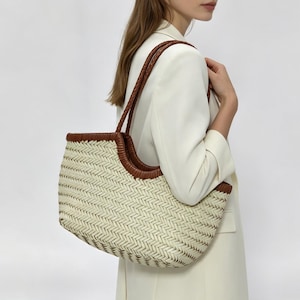 Könnte beinhalten: Eine geflochtene Tragetasche mit cremefarbenem Körper im Fischgrätenmuster und braunem Lederbesatz entlang der Oberkante und der Griffe. Die Tasche hat ein breites, offenes Design mit zwei geflochtenen Schulterriemen aus Leder.