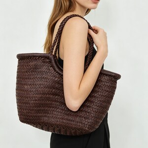Puede incluir: Un bolso tote de cuero trenzado en color marrón oscuro con un diseño abierto, una base redondeada estructurada y dos asas de hombro trenzadas. El exterior muestra un patrón de tejido en espiga uniforme en toda la superficie.