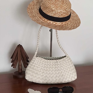 Può includere: Un cappello di paglia con nastro nero, una borsa a spalla intrecciata bianca con manici intrecciati, un piccolo oggetto in legno a forma di albero, una sagoma bianca a forma di farfalla e occhiali da sole ovali neri su una superficie.
