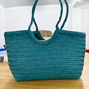Könnte beinhalten: Eine blaugrüne Handtasche mit einem geflochtenen Fischgrätenmuster über den gesamten Körper. Die Tasche hat zwei geflochtene Henkel, die an der Oberkante befestigt sind, welche sich in der Mitte nach unten wölbt. Die Form ist rechteckig.