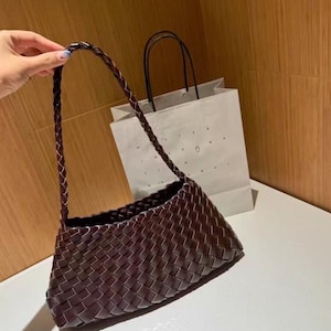 Peut inclure: Un sac à main en cuir tressé marron foncé doté d'un corps rectangulaire et d'une bandoulière tressée. Le sac présente un motif entrelacé distinct sur toute sa surface. Derrière le sac se trouve un sac en papier blanc avec du texte.