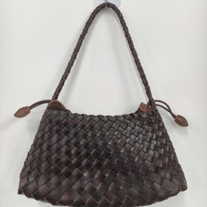 May include: A dark brown handbag featuring a woven basket-weave pattern across the entire body. The bag includes a single braided shoulder strap and two side drawstrings with small, pointed brown tabs at the ends for closure adjustment.