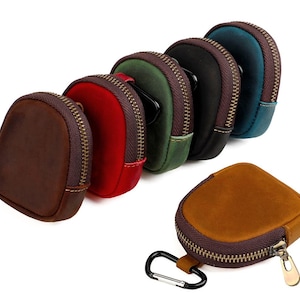 May include: A collection of small, round leather pouches in various colors, including brown, red, green, black, and teal. Each pouch has a contrasting brown zipper and a metal zipper pull. One pouch has a carabiner clip attached.