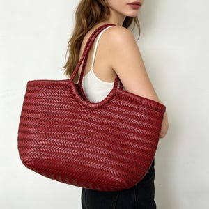 May include: A deep red tote bag featuring a woven herringbone pattern throughout the exterior. The bag has two integrated shoulder straps and a wide, open-top design, constructed with a consistent braided texture across the entire surface.