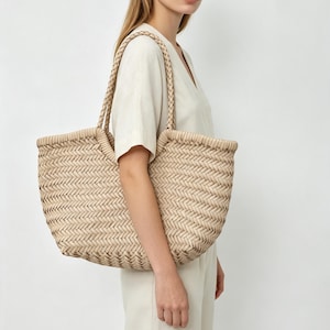 Könnte beinhalten: Eine große beige Tragetasche mit einem geflochtenen Fischgrätenmuster über den gesamten Körper. Die Tasche verfügt über zwei geflochtene Schulterriemen und eine geschwungene Öffnung, gefertigt aus einem hellen Material mit gleichmäßiger Textur.