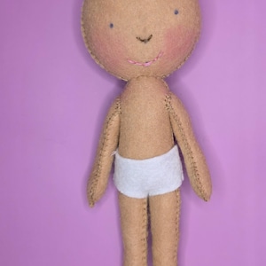 Puede incluir: Muñeca hecha a mano con cuerpo color canela, rasgos cosidos y una cara sonriente con mejillas rosadas y boca cosida rosa. La muñeca lleva un pantalón corto blanco y tiene extremidades cosidas. El fondo es morado liso.