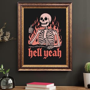 May include: Framed art print featuring a skeleton giving a thumbs up, surrounded by flames. The words "hell yeah" are written in red gothic font below the skeleton. The artwork is set against a black background, framed in a brown wooden frame.