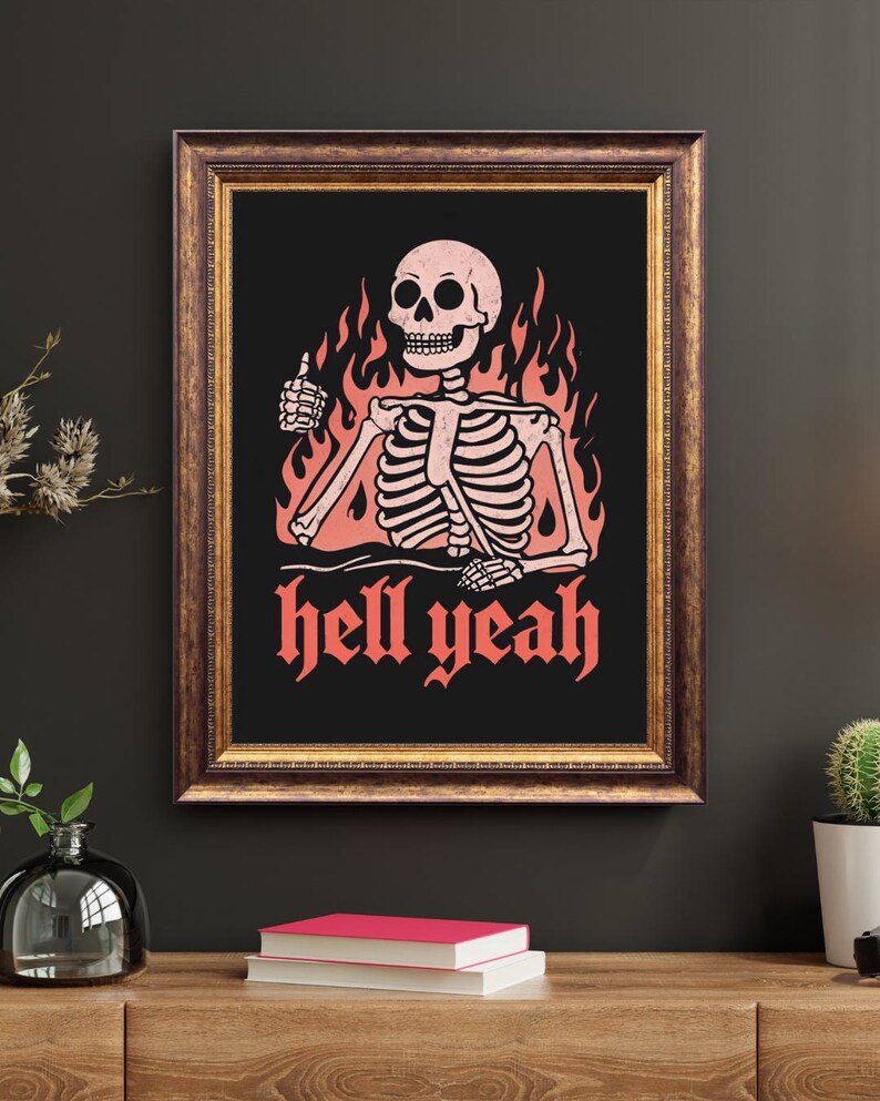May include: Framed art print featuring a skeleton giving a thumbs up, surrounded by flames. The words "hell yeah" are written in red gothic font below the skeleton. The artwork is set against a black background, framed in a brown wooden frame.