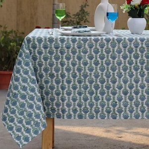 May include: A rectangular tablecloth with a repeating blue floral pattern and green leaves. The table is set with white plates, glasses with green and blue liquid, and white vases with flowers and decorative stems. The tablecloth is draped over a wooden table.