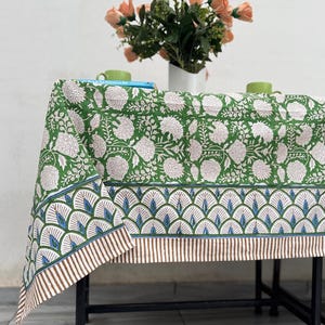 May include: A green tablecloth with a floral pattern and a decorative border. The tablecloth is draped over a black metal table. A vase of peach-colored flowers sits on the table, along with two green mugs.