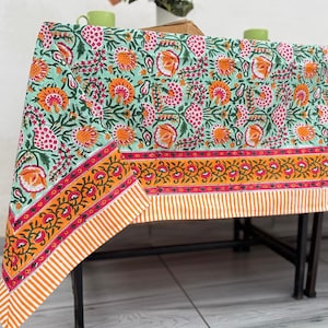 Puede incluir: Un mantel rectangular con un estampado floral en tonos turquesa, naranja, rosa y blanco. El borde presenta un diseño naranja y rojo con un borde a rayas. Un jarrón con flores color melocotón está sobre la mesa.
