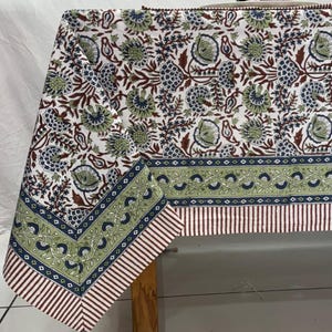 May include: A rectangular tablecloth featuring a white base with a repeating floral pattern in shades of green, blue, and brown. The border includes a green floral band, a thin blue geometric line, and a striped edge in brown and white.