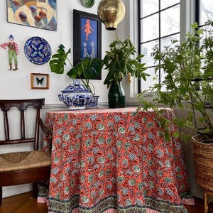 May include: A room with a round table draped in a red floral tablecloth. The room is decorated with artwork, a blue and white ceramic serving dish, and a green vase with leafy branches. A wooden chair is in the foreground.