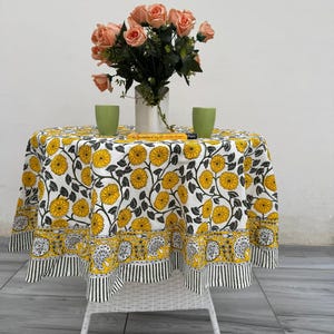 May include: A round table with a white wicker base is covered with a floral tablecloth featuring yellow flowers and black leaves on a white background. A vase of peach roses and two green cups sit on the table.