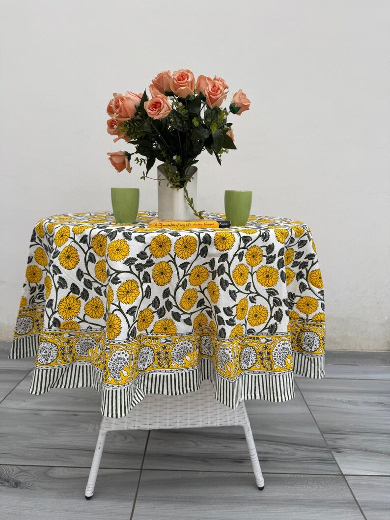 May include: A round table with a white wicker base is covered with a floral tablecloth featuring yellow flowers and black leaves on a white background. A vase of peach roses and two green cups sit on the table.