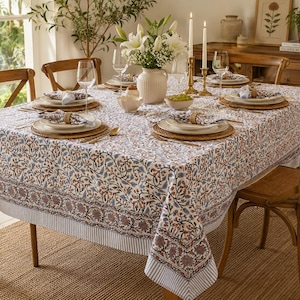 May include: A rectangular tablecloth featuring a detailed floral pattern in shades of blue, brown, and orange on a white background, finished with a striped border. The fabric drapes over a dining table set with white plates, glassware, and napkins.