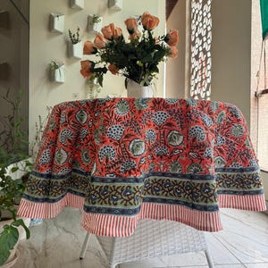 May include: Round tablecloth with a floral pattern in shades of red, blue, and green. The tablecloth is draped over a white wicker chair. A vase of orange roses sits on the table.