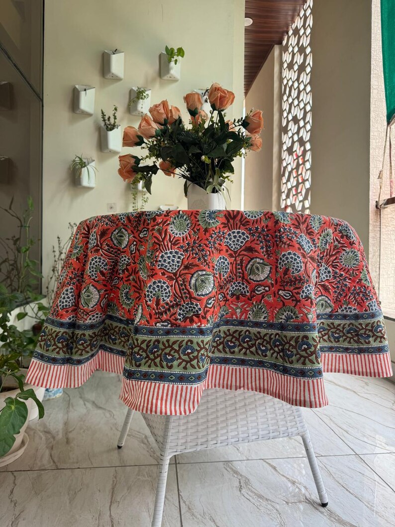May include: Round tablecloth with a floral pattern in shades of red, blue, and green. The tablecloth is draped over a white wicker chair. A vase of orange roses sits on the table.