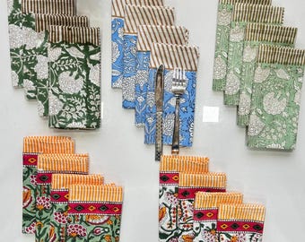 May include: A selection of patterned cloth napkins in various colours. The napkins feature floral and striped designs in green, blue, orange, and white. Some napkins are folded with a knife and fork inside.