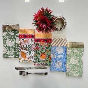 May include: A collection of five colorful floral print cloth napkins. Each napkin features a different color scheme, including green, white, red, orange, and blue. A knife and fork are placed near the napkins. A red flower and a decorative candle holder are also in the image.