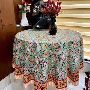 Op de afbeelding: Een ronde tafelkleed met een bloemenpatroon in groen, roze, geel en oranje tinten. Het tafelkleed is over een tafel gedrapeerd, met een decoratieve rand en een streepjespatroon. Een zwarte gitaar en een vaas met bloemen staan op de achtergrond.