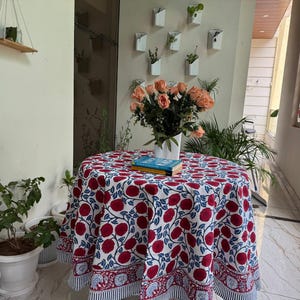 May include: A round tablecloth with a red floral pattern on a white background. The tablecloth is draped over a small table, with a vase of peach roses and a stack of books on top. The table is surrounded by potted plants.
