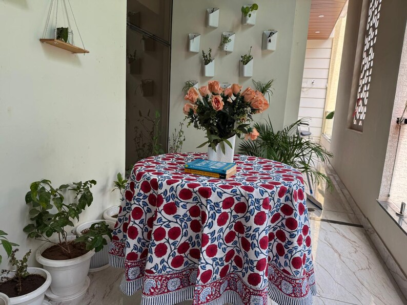 May include: A round tablecloth with a red floral pattern on a white background. The tablecloth is draped over a small table, with a vase of peach roses and a stack of books on top. The table is surrounded by potted plants.