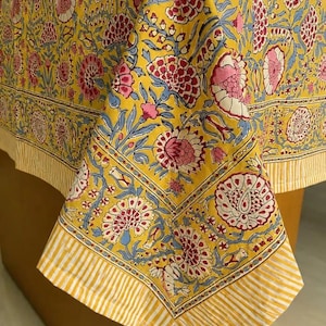 May include: A yellow tablecloth featuring a repeating floral pattern in shades of pink, red, and blue. The fabric displays a central block-printed botanical design bordered by a distinct yellow and white striped edge along the bottom hem.