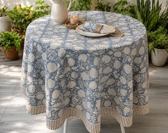 Può includere: Una tovaglia rotonda caratterizzata da un motivo floreale stampato in blu e bianco con un bordo a righe. Il tessuto copre un tavolo circolare, mostrando intricati motivi botanici e una palette di colori coerente in tutto il design tessile.