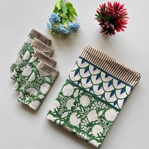 May include: A green and white floral patterned tablecloth and matching napkins. The tablecloth has a brown striped border and a blue and white scalloped design. The napkins have a brown striped border. Decorative flowers are in the background.