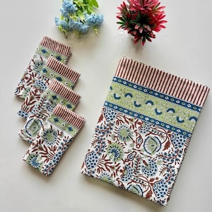 Op de afbeelding: Een witte tafelkleed en bijpassende servetten met een bloemenpatroon in blauw, groen en bruin. De servetten hebben een gestreepte rand. Decoratieve bloemen in blauw en rood staan op de achtergrond.