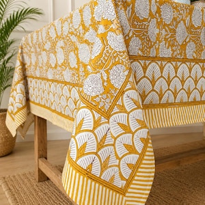 May include: A rectangular tablecloth featuring a mustard yellow and white colour scheme. The fabric displays a detailed floral pattern on the top surface, bordered by a repeating fan-shaped motif and a striped edge along the draped corners.