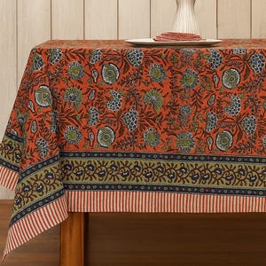 Può includere: Una tovaglia rettangolare arancione con un motivo floreale verde, blu e bianco. Il bordo presenta un motivo floreale verde e blu sopra strisce rosse e bianche. Un vaso di fiori è sul tavolo.