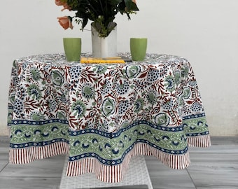 Può includere: Un tavolo rotondo con una tovaglia floreale verde, blu e marrone. Il tavolo è apparecchiato con un bouquet di rose color pesca, due bicchieri verdi e un libro. Il tavolo è sostenuto da una base in vimini bianca. La tovaglia ha un bordo fantasia.