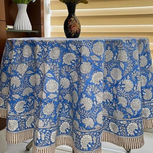 Può includere: Una tovaglia rotonda blu con un motivo floreale bianco. La tovaglia ha un bordo a righe marrone e bianco. Un vaso con fiori è sul tavolo. La tovaglia ha un diametro di circa 152 cm.
