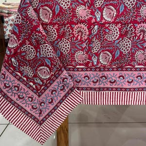 Può includere: Una tovaglia rossa con un motivo floreale bianco, rosa e blu. La tovaglia ha un bordo a righe rosse e bianche. Il tessuto è drappeggiato su un tavolo di legno.