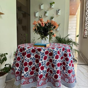 Puede incluir: Un mantel redondo con un estampado floral en rojo y azul sobre un fondo blanco. El mantel está sobre una pequeña mesa blanca. Un jarrón con rosas naranjas está sobre la mesa, con libros debajo.