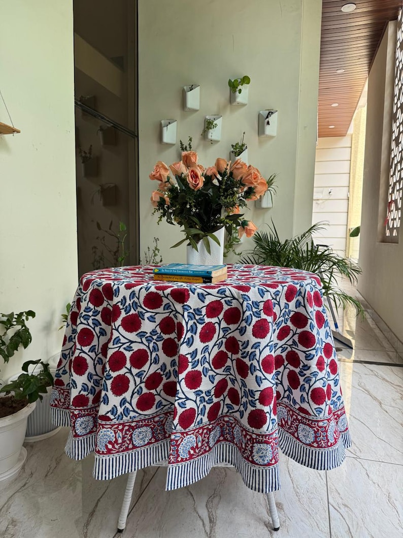May include: A round tablecloth featuring a floral design in red and blue on a white backdrop. The cloth covers a small white table. A vase of orange roses sits on the table, with books underneath.