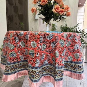 May include: A round coral tablecloth with a floral pattern in blue, green, and brown. The border features a striped design in red and white. A bouquet of peach roses sits on the table.