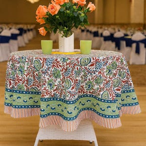 May include: A round table with a floral tablecloth. The tablecloth features a white background with red, blue, and green floral patterns. A vase of orange roses and two green glasses are on the table.