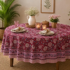 May include: A round tablecloth featuring a deep red base with a repeating floral and paisley pattern in shades of pink, white, and blue. The fabric has a wide border with a striped edge, draped over a circular table set with plates and glasses.