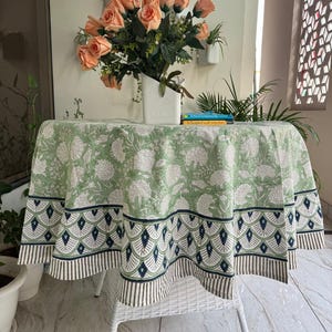 May include: A round tablecloth with a green and white floral pattern, featuring a navy and white border. The cloth covers a white table. A vase of peach roses sits on the table. The table is approximately 120 cm in diameter.
