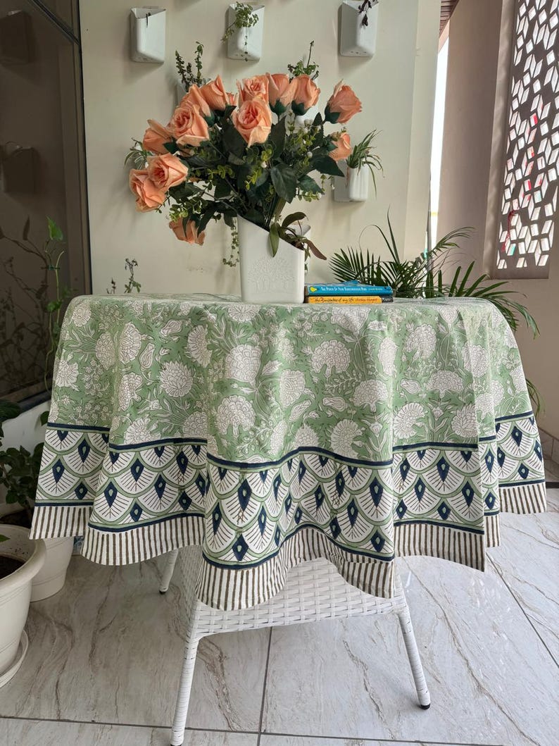 May include: A round tablecloth with a green and white floral pattern, featuring a navy and white border. The cloth covers a white table. A vase of peach roses sits on the table. The table is approximately 120 cm in diameter.