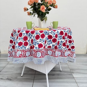 May include: A round tablecloth with a floral pattern in red and blue on a white background. The tablecloth is on a small white table with two green mugs and a vase of peach roses. The table is on a tiled patio.