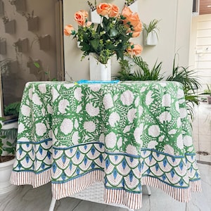 May include: A round green tablecloth with a white floral pattern covers a white table. The tablecloth has a blue, green, and brown scalloped border. A vase of peach-colored roses sits on the table.