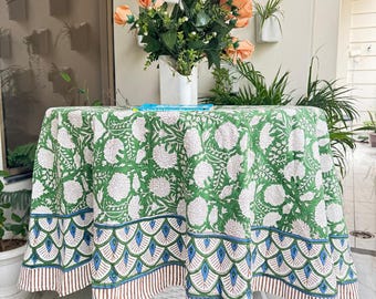 Può includere: Una tovaglia rotonda verde con un motivo floreale bianco copre un tavolo bianco. La tovaglia ha un bordo a conchiglia blu, verde e marrone. Un vaso di rose color pesca è sul tavolo.